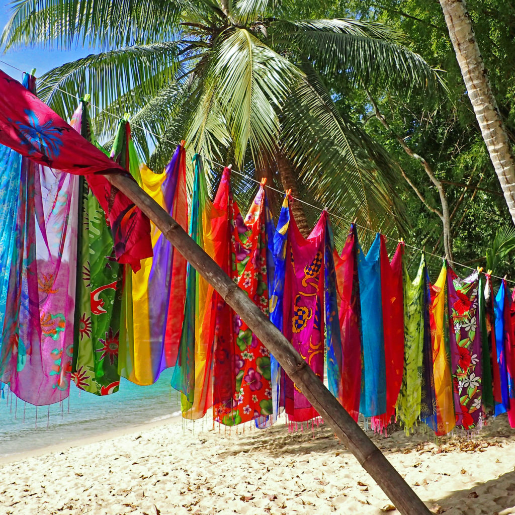 Colorful fabric scarves or cloth sarongs printed with beautiful tropical designs hanging in a line and held up by a bamboo pole over a sunny palm tree lined beach in front of calm aquamarine water.